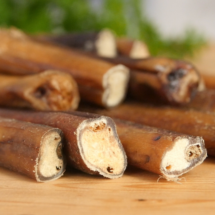 Close-up of several bully sticks showing their textured brown outer skin and pale beige inner marrow, arranged on a wooden surface with a blurred green background.